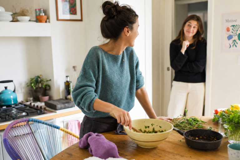 Vrouw roert in een kom aan de keukentafel terwijl een andere vrouw glimlachend in de deuropening staat.